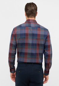 Uomo di spalle che indossa una camicia a maniche lunghe a quadri nei toni del blu, rosso e grigio, infilata in pantaloni scuri blu navy.