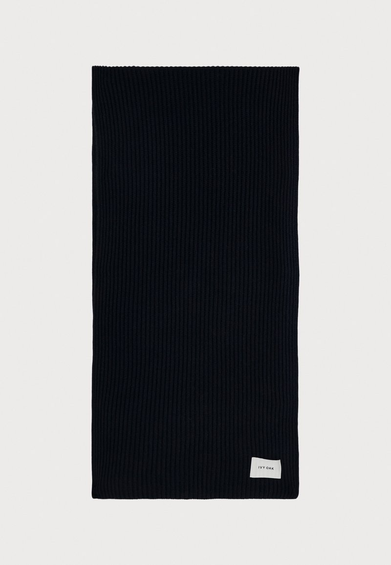 Ribbede strikscarf i mørkeblå, rektangulær form, med et lille hvidt mærke med logo i den ene ende. Blødt materiale, alsidigt design.