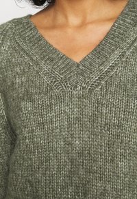 Oliwkowy sweter wykonany na drutach ma dekolt w kształcie litery V, prążkowaną strukturę i luźny krój. Materiał wydaje się miękki z różnymi grubościami.