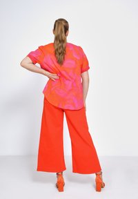 Heldere oranje wijde broek in combinatie met een levendig roze en oranje geblokte blouse. De outfit heeft korte mouwen en een gebogen zoom.