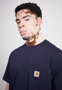 Tengerészkék, rövid ujjú pamut póló, mellzsebbe varrt Carhartt logós felvarróval. Sima textúra, minimalista kialakítás.