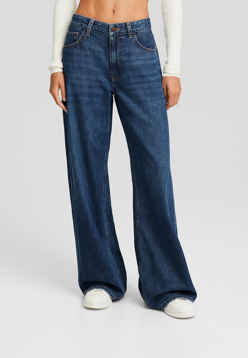 Weite Jeans aus dunkelblauem Denim mit subtilen Verblassungen, einem Knopfverschluss und Gürtelschlaufen. Kombiniert mit weißen Sneakern.