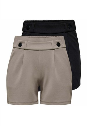 Junge Frau steht mit einer Hand in der Tasche der schwarzen Shorts, trägt ein weißes Kurzarmoberteil mit großem schwarzem Blumendesign und schwarze Ballerinas.