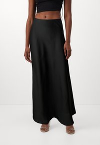 Jupe maxi en satin noir avec une texture lisse et un design fluide. Silhouette épurée s'étendant jusqu'aux chevilles, associée à des sandales à talons transparents.