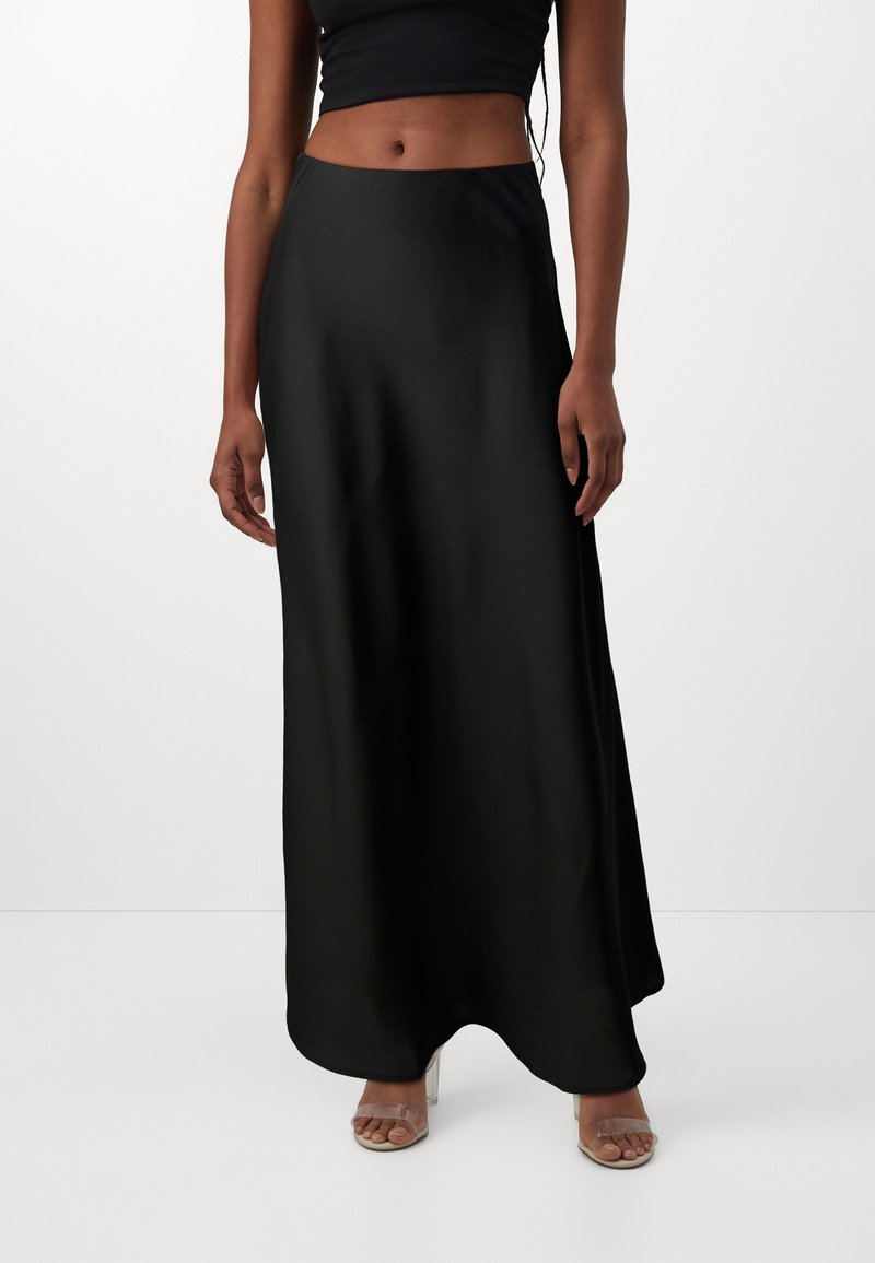 Jupe maxi en satin noir avec une texture lisse et un design fluide. Silhouette épurée s'étendant jusqu'aux chevilles, associée à des sandales à talons transparents.