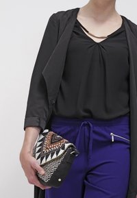 Svart blus med rynkad halsringning, grå yttre lager och blåa dragsko-byxor. Håller en mönstrad kuvertväska med geometriska mönster.