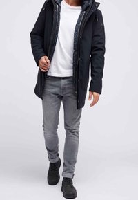 Homme marchant portant une veste à capuche noire, un t-shirt blanc, un jean gris et des baskets noires sur un fond clair uni.