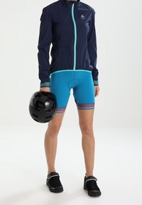 Coupe-vent marine avec fermeture éclair turquoise, short bleu avec accents rayés, casque noir à la main et chaussures de sport noires.