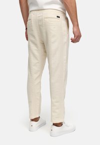 Hellbeige Baumwollhosen mit einem strukturierten vertikalen Muster, ausgestattet mit zwei Gesäßtaschen und einem lässigen Schnitt. Kombiniert mit weißen Sneakers.