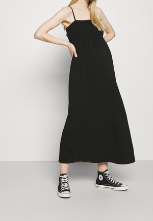 Mujer con un vestido maxi negro sin mangas con tirantes de espagueti combinado con zapatillas negras altas, de pie con las manos en las caderas.