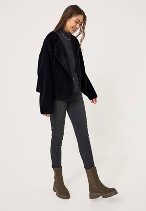 Giacca in pile nera con maniche larghe, design a strati, indossata sopra un dolcevita nero. Jeans neri a vestibilità slim e stivali alla caviglia marroni completano il look.