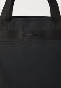 Sac fourre-tout en tissu noir avec des anses robustes, comportant un patch logo et une surface lisse et texturée. Design simple et fonctionnel.