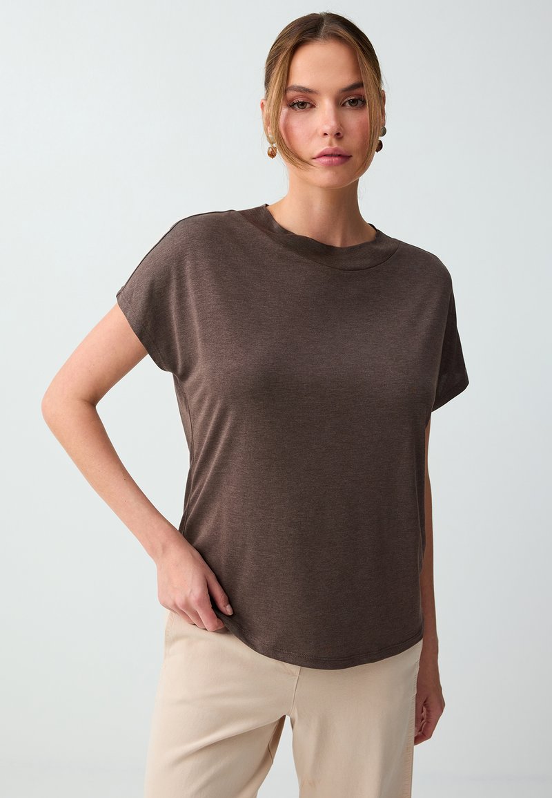 Femme aux cheveux châtain clair, portant un haut marron à manches courtes et un pantalon beige, debout devant un fond gris clair uni.
