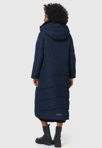 Long navy blue puffer coat with a hood, vertical quilted design, and contrasting black accents. Features side pockets and a brand label.