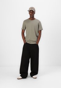 Olivenfarbener Baumwoll-T-Shirt, lockere Passform, kombiniert mit weiten schwarzen Hosen. Das Schuhwerk hat eine weiße Basis und beige Akzente. Eine Fußballmütze auf dem Kopf.