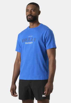 Hombre con camiseta azul de Helly Hansen y pantalones cortos negros, de pie contra un fondo claro y liso, mirando ligeramente hacia un lado.