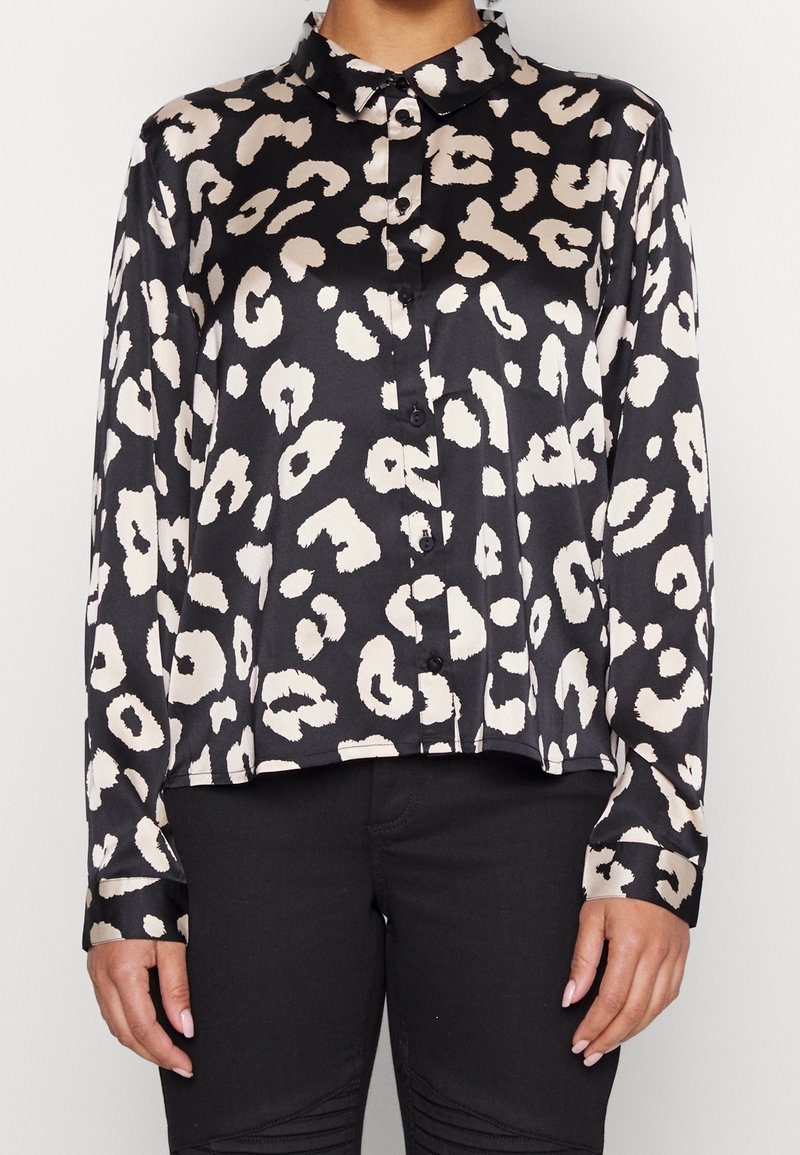 Blouse noire à boutons avec des taches léopard blanches irrégulières portée avec un pantalon noir, vue de face du cou aux hanches.