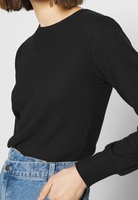 Maglione nero a costine con maniche lunghe, scollatura rotonda, forma aderente e dettagli in tessuto testurizzato, abbinato a jeans di denim chiaro.