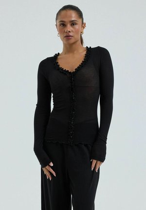 Femme aux cheveux tirés en arrière portant un cardigan noir cintré à manches longues et un pantalon noir, debout devant un fond uni clair.