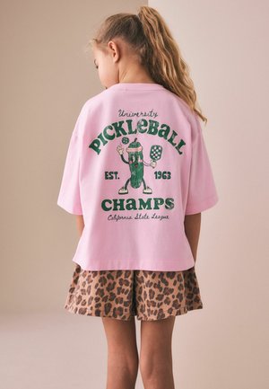 Young girl with long hair in a ponytail wearing a pink oversized t-shirt with a pickleball design and leopard print shorts, facing away.