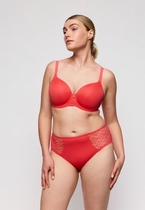 Vrouw met blond haar in een lage paardenstaart, gekleed in een rode beha en bijpassende hoog uitgesneden slip met kant, staand tegen een effen witte achtergrond.