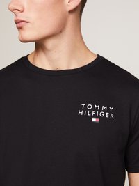 Zwarte katoenen t-shirt met witte "TOMMY HILFIGER" tekst en een klein logo met rode, witte en blauwe accenten op de linkerborst.