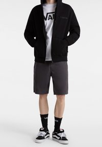 Person trägt schwarzen Kapuzenpullover mit Reißverschluss, weißes Vans-T-Shirt, graue Shorts, schwarze Vans-Socken und schwarz-weiße Vans-Sneaker, steht mit den Händen in den Taschen.