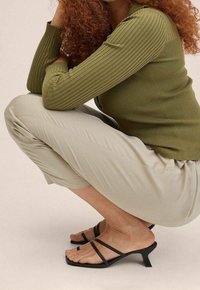 Personne aux cheveux roux bouclés accroupie, vêtue d’un haut côtelé vert olive, d’un pantalon beige clair et de sandales noires à talons à brides, sur un fond uni.