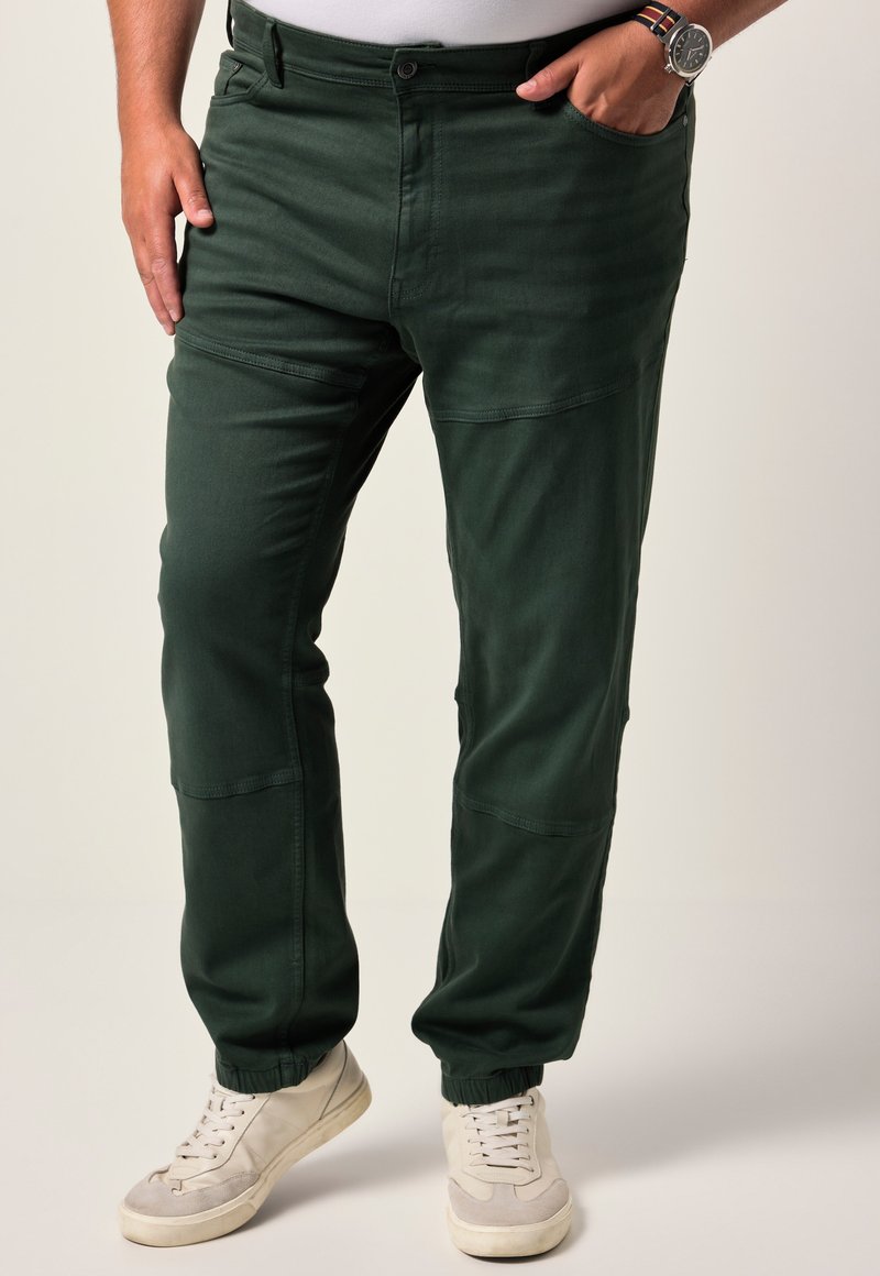 Hombre con pantalones verdes oscuros ajustados en el tobillo con puños elásticos y zapatillas blancas, mano en el bolsillo y reloj en la muñeca.