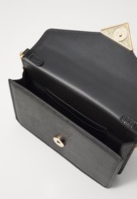 Sac à main noir avec un extérieur texturé, quincaillerie dorée et un accent triangulaire ; dispose d'un intérieur ouvert avec des fentes pour cartes et une fermeture magnétique.