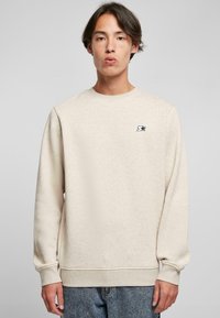 Sudadera beige hecha de una tela suave con mangas largas y cuello redondo. Presenta un pequeño logo negro en el pecho y puños de canalé.
