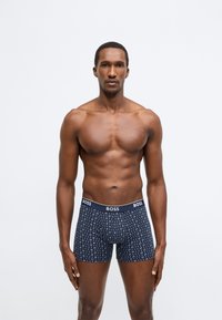 Sötétkék rövidnadrág jellegű alsónadrág márkázott derékpánttal. Világosabb színű geometriai mintázattal. Az anyag simának és rugalmasnak tűnik.