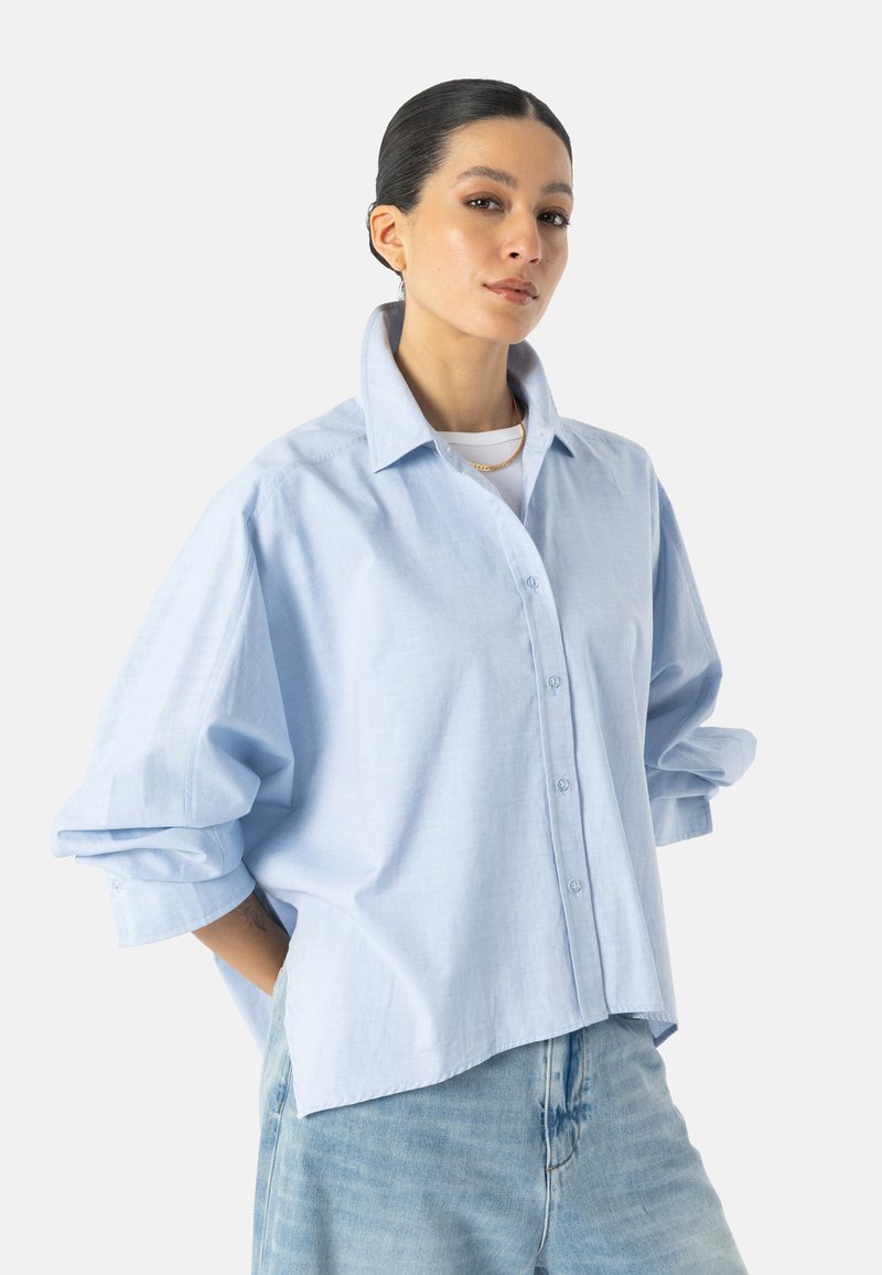 Femme aux cheveux foncés attachés en chignon bas, portant une chemise oversize bleu clair boutonnée sur un haut blanc et un jean bleu clair, debout les mains dans les poches.