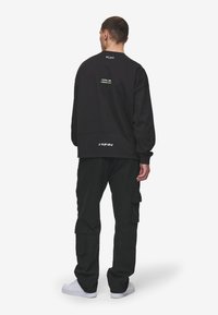 Mustapohjainen crewneck-huppari, jossa on painettu teksti selässä ja logo helmassa, yhdistettynä mustiin cargo-housuihin. Malli seisoo selkä käännettynä.