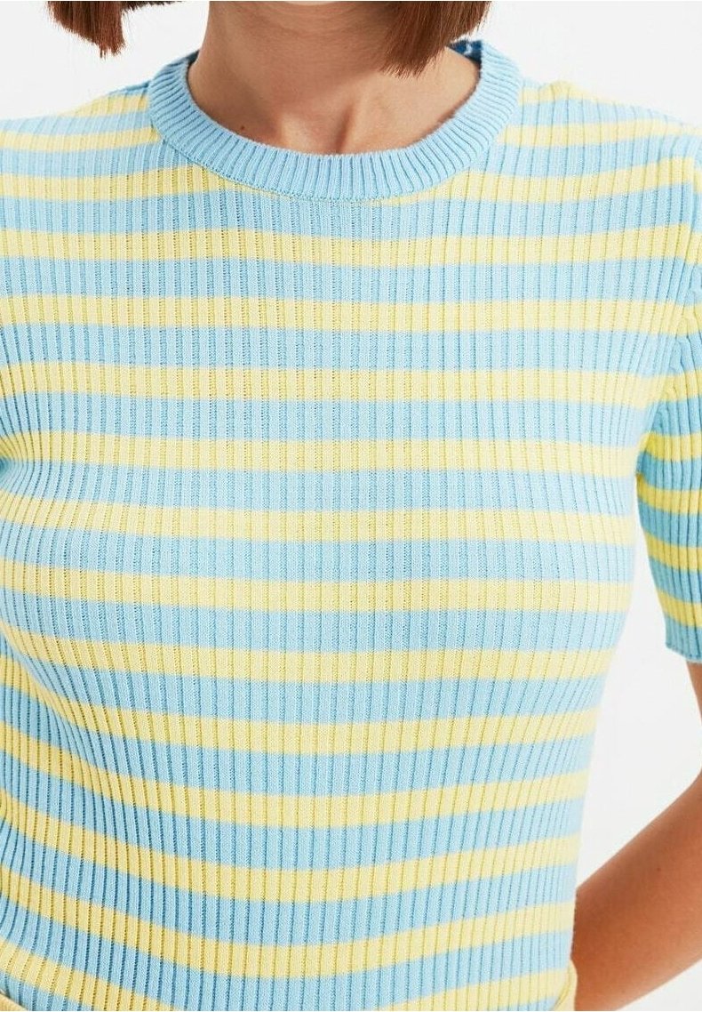 Femme portant un pull en maille côtelée avec des rayures horizontales bleues et jaunes et un col rond, manches courtes visibles sur fond blanc.