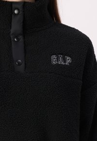 Zwarte fleece trui met hoge kraag, drukknoopsluiting en geborduurd "GAP" logo. Zacht van textuur met een pluizige, warme uitstraling.