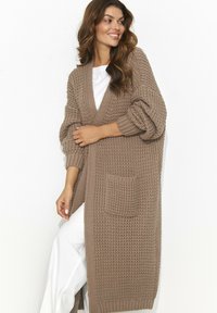 Sweter z dzianiny w jasnobrązowym kolorze, charakteryzujący się luźnym krojem, głębokim dekoltem w kształcie litery V, prążkowanymi mankietami oraz kieszenią z przodu. Tekstura jest gruba, z tkanym wzorem.