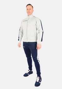 Giacca grigia chiaro con zip e pannelli laterali blu, abbinata a pantaloni blu con strisce bianche. Sneakers bianche e nere completano l'outfit.