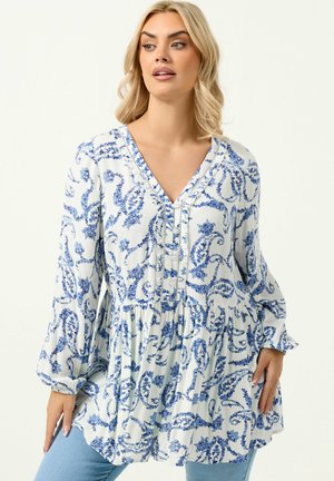 Lyshåret kvinde iført en hvid bluse med blå blomster- og paisleymønstre og lange ærmer, kombineret med lyseblå jeans.