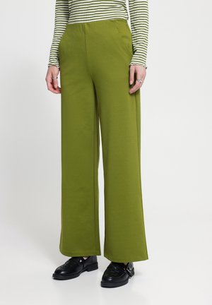 Personne portant un pantalon vert à jambes larges, des mocassins noirs et un haut blanc à manches longues avec des rayures vertes, debout devant un fond uni.
