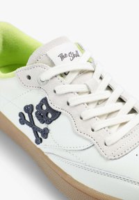 Sneaker blanche avec des accents en suède gris, dotée d'une doublure intérieure verte, de lacets blancs et d'un design brodé de crâne et os croisés.