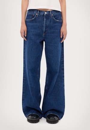 Person trägt dunkelblaue Jeans mit weitem Bein und schwarze klobige Stiefel, steht vor einem schlichten hellen Hintergrund.