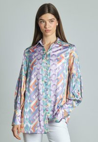 Camicia con bottoni in tessuto iridescente che presenta motivi geometrici in lavanda, teal, arancione e bianco, con maniche lunghe a sbuffo.
