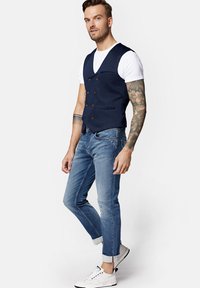 Granatowa kamizelka dwurzędowa, biała koszulka pod spodem, w połączeniu z jasnoniebieskimi spodnimi slim-fit i białymi trampkami z czarnymi akcentami.