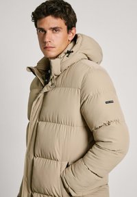 Beige puffer jacket with a hood, featuring quilted sections, a front zipper, and logo detail on the left sleeve. Soft texture.