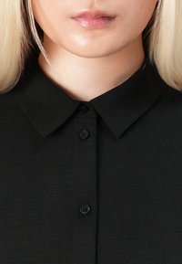 Close-up of a person’s lower face and neck wearing a black buttoned shirt with a folded collar and straight blonde hair.