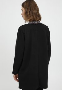 Cappotto nero texture con maniche lunghe e orlo curvo. Presenta un colletto a contrasto con motivo. La vista posteriore mostra linee semplici e pulite.