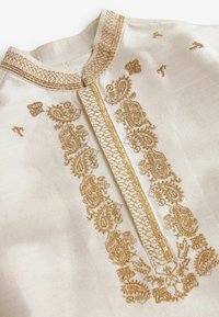 Tissu blanc avec une broderie dorée détaillée en motifs cachemire le long d'une couture verticale et du col, mettant en valeur des points décoratifs complexes.