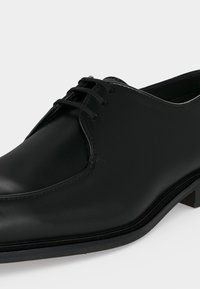 Μαύρο επίσημο δερμάτινο παπούτσι με κορδόνια, στρογγυλεμένη μύτη και χαμηλό τακούνι, απεικονίζεται σε πλευρική όψη σε λευκό φόντο.