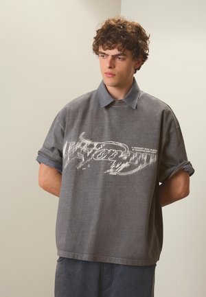 Junger Mann mit lockigem Haar, der ein graues übergroßes T-Shirt mit weißem grafischem Text trägt, das über einem gemusterten Hemd mit Kragen liegt, Hände hinter dem Rücken.